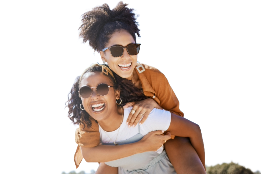 two people wearing sunglasses with one giving a piggyback ride to the other smile against a blue sky backdrop